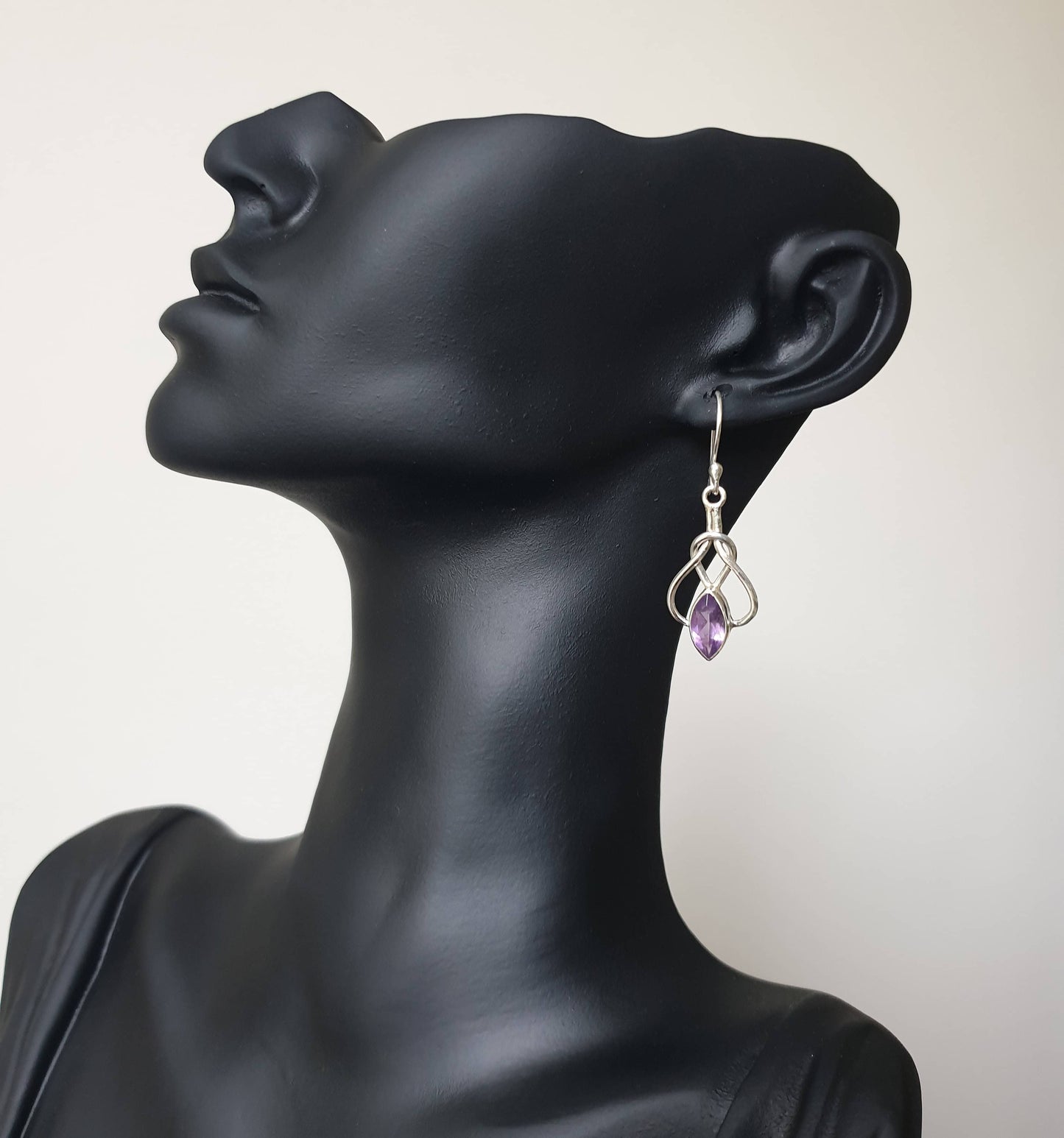 This image displays a pair of elegant Amethyst earrings, presented on a mannequin head to highlight their design and flow. The earrings are crafted with intricate detailing, featuring a small purple amethyst stone at the center, surrounded by a loop of delicate silver wiring that hangs gracefully from the ear.