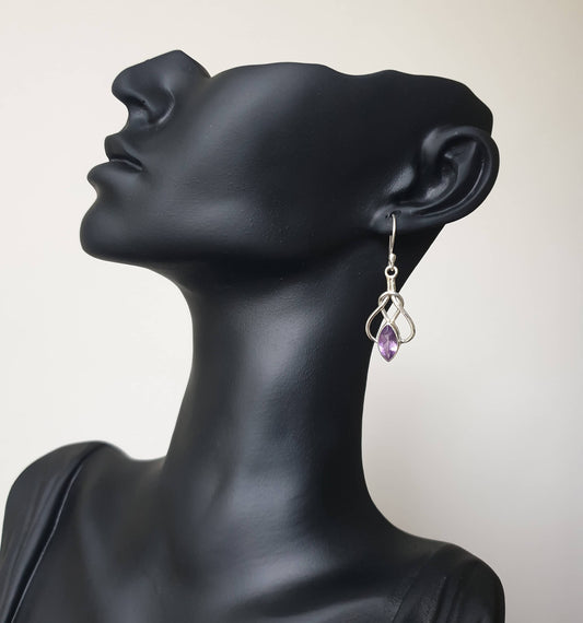 This image displays a pair of elegant Amethyst earrings, presented on a mannequin head to highlight their design and flow. The earrings are crafted with intricate detailing, featuring a small purple amethyst stone at the center, surrounded by a loop of delicate silver wiring that hangs gracefully from the ear.