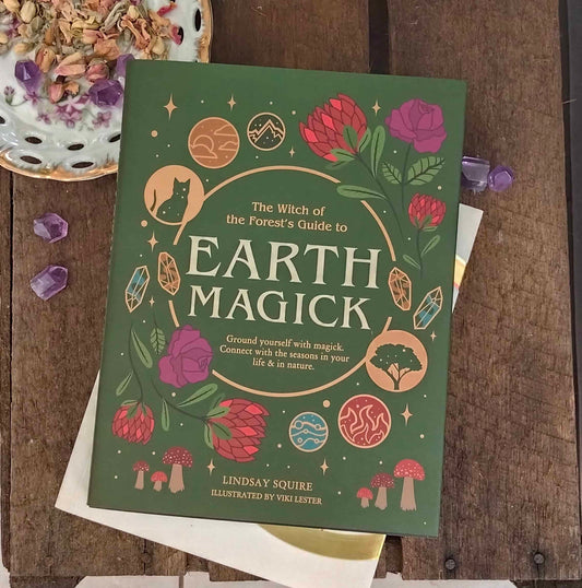Book titled 'The Witch of the Forest's Guide to Earth Magick' on a wooden surface with decorative elements.