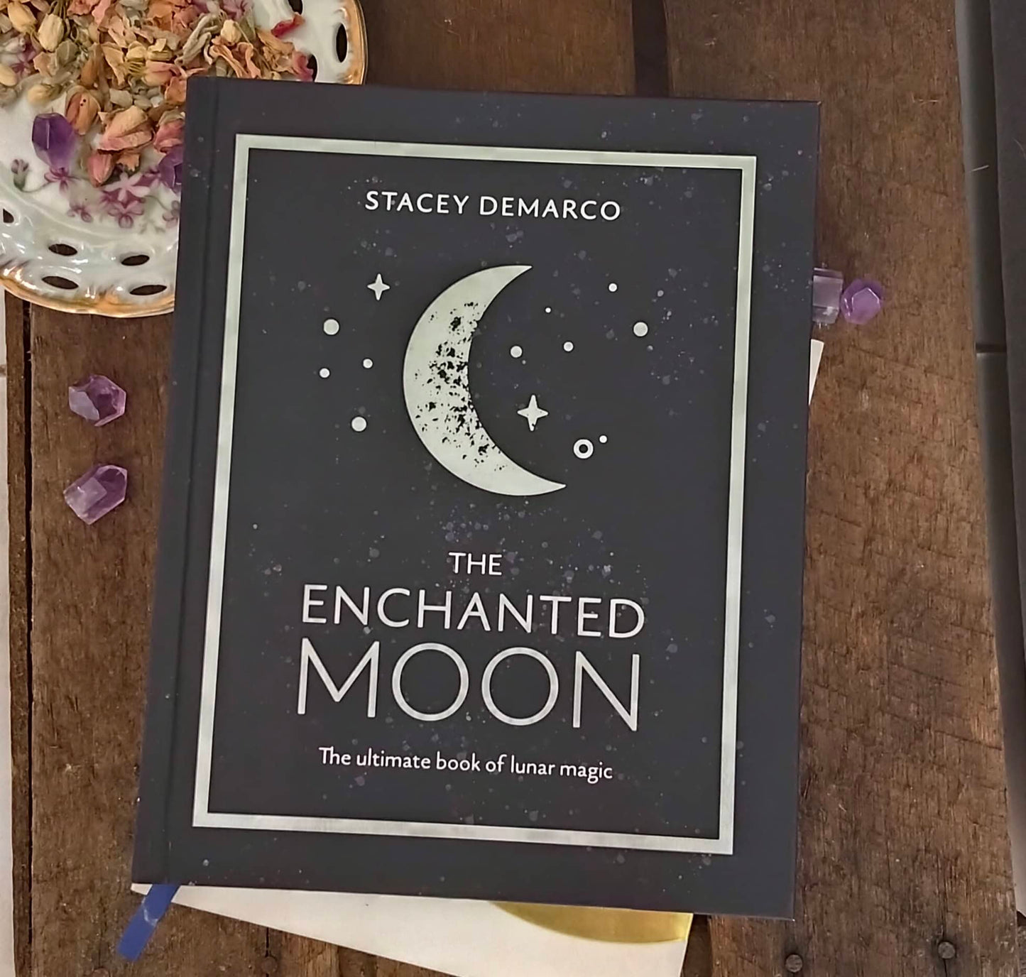 Book titled 'The Enchanted Moon' by Stacey Demarco on a wooden surface with decorative elements.